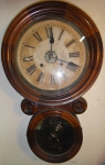 Relógio de parede, com despertador, da marca "Waterbury". Caixa em madeira clara em feitio de "8". E.U.A. - 1880. Funcionando. (Mostrador com pequenas manchas).