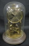 Antigo relógio alemão para cima de móvel com corda para 360 dias. Estrutura e mostrador em metal dourado. Base em madeira. Cúpula em vidro moldado. Funcionando.