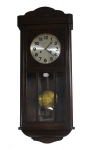 Relógio de parede da marca "Junghans". Caixa em madeira clara com porta envidraçada. Frontão ondulado e entalhado com florão. Alt.: 80cm. Comp.: 32cm. Funcionando.