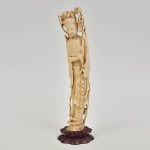 Divindade "Kuan Yin com abano e ramos de peônias" esculpida em marfim. Base em madeira no feitio de flor de lótus. Alt.: 33cm. China - 1940.